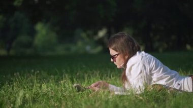 Güneş gözlüklü esmer kadın kitap okuyor ve etrafa bakıyor.