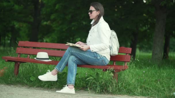 Young woman brunette sits on red wooden bench in park — Stock Video