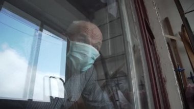Mavi maskeli emekli adam eski pencereden dışarı bakıyor.