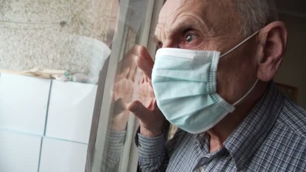 Oudere gepensioneerde met gezichtsmasker kijkt door verouderd raam — Stockvideo