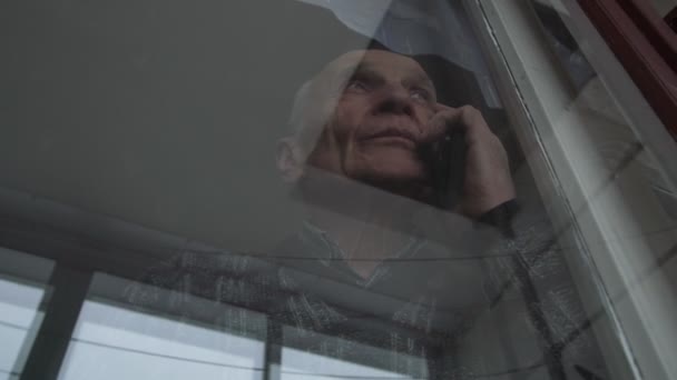 Gubben pratar på smartphone tittar utanför balkongen fönster — Stockvideo