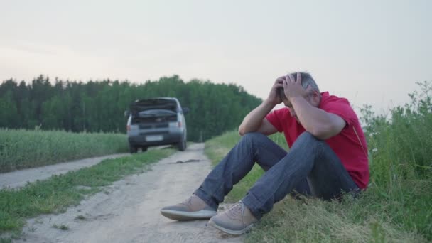 Uomo sconvolto con i capelli grigi si siede sul lato della strada sterrata in campo vicino a un'auto rotta — Video Stock