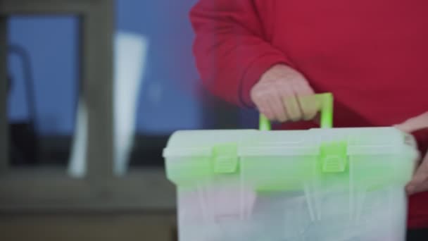 Старый ремонтник в красном свитере кладет пластиковый ящик на стол — стоковое видео