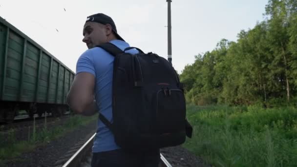 Viajero con mochila camina a lo largo de carriles de ferrocarril cerca de tren de paso — Vídeo de stock