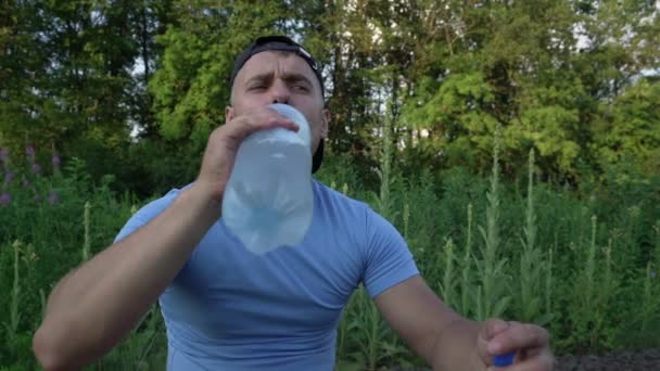 Şapkalı ve mavi gömlekli adam oturur ve şişeden su içer. — Stok video