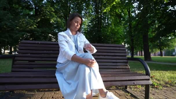 Middle aged woman in white jacket looks at watch on hand in park — Stock Video