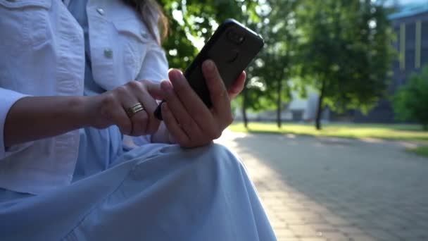 Señora de chaqueta blanca rojo sms en el teléfono sentado en el parque — Vídeos de Stock