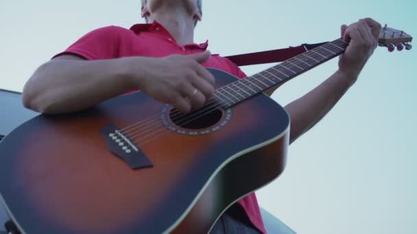 Mann im roten Trikot spielt Akustikgitarre vor strahlend blauem Himmel — Stockvideo