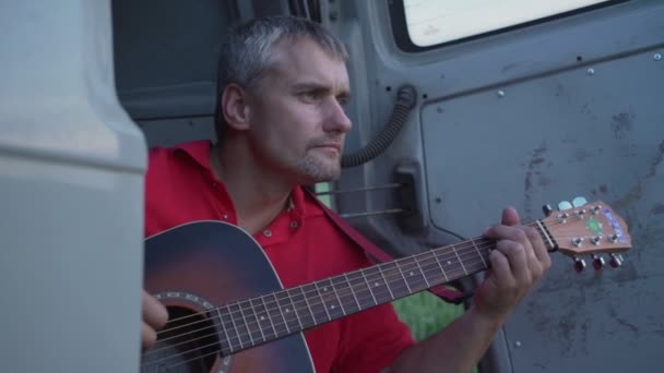 Уставший мужчина играет на акустической гитаре, сидя на краю минивэна — стоковое видео