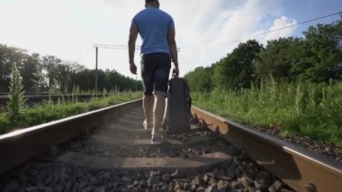 Şortlu üzgün adam demiryolu boyunca yürüyor ve yol çantası taşıyor.