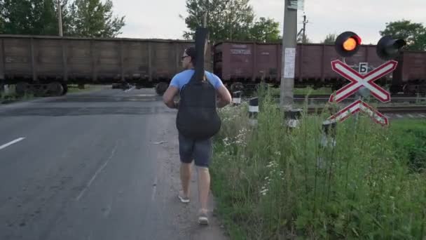 Hombre con guitarra se acerca al cruce ferroviario a lo largo del cual circula el tren de mercancías — Vídeo de stock