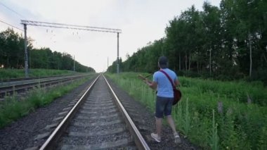 Şapkalı ve şortlu müzisyen tren yolunda yürüyor ve gitar çalıyor.