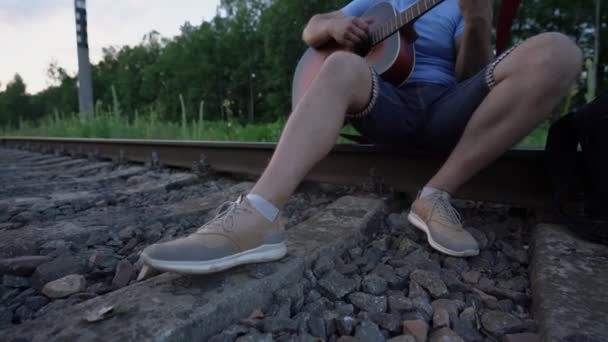 Viaggiatore maschio si sedette a riposare su rotaia della ferrovia mettere zaino e suona la chitarra — Video Stock