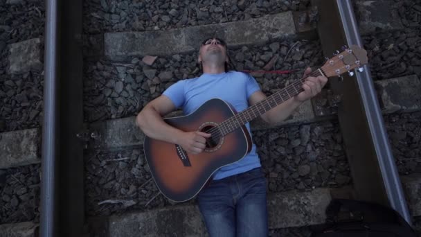 Hombre en gafas de sol se encuentra en traviesas de ferrocarril toca la guitarra y canta — Vídeo de stock