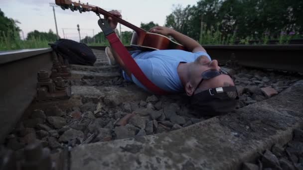 Mannelijke muzikant met baard in zonnebril alleen ligt op dwarsliggers van de spoorweg — Stockvideo