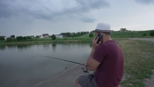 Rybář v klobouku sedí s rybářským prutem na židli a mluví telefonicky — Stock video