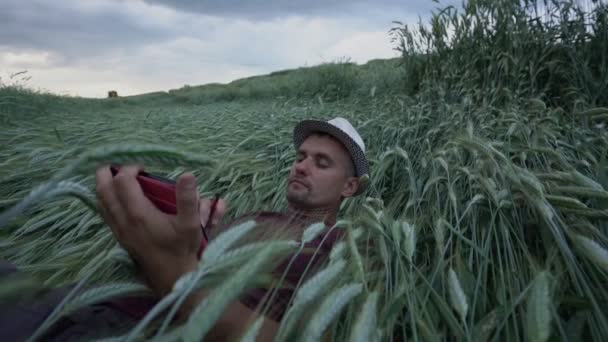 Man in hoed met baard ligt op tarweveld en werkt in tablet — Stockvideo