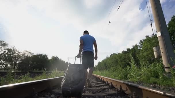 Şortlu yorgun adam tren yolunda yürüyor ve yol çantasını çekiyor. — Stok video