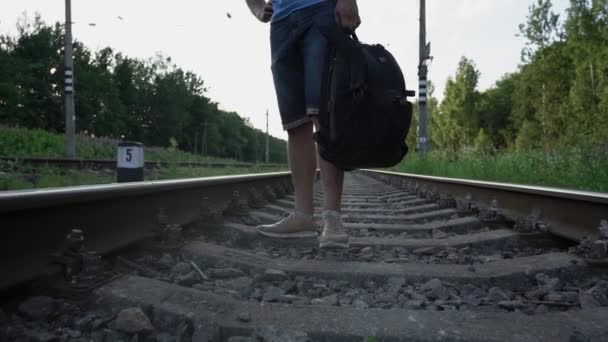 Homem caminha lentamente ao longo de trilhos de trem e carrega mochila em suas mãos — Vídeo de Stock