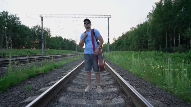 Homem sério em óculos de sol e passeios de boné ao longo de trilhos de trem — Vídeo de Stock
