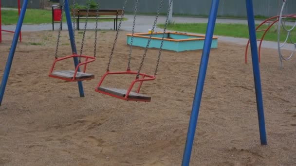 El viento balancea los columpios rojos en las cadenas en el parque infantil vacío — Vídeo de stock