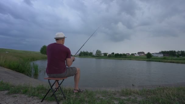 Man vissen zittend op stoel op oever van vijver in het zicht van achteren — Stockvideo