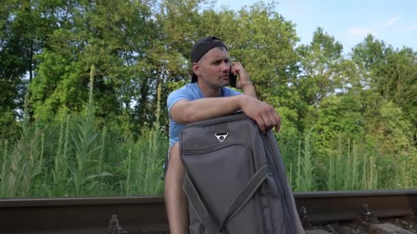 Un hombre con una maleta sentada sobre raíles en el ferrocarril hablando por teléfono — Vídeos de Stock