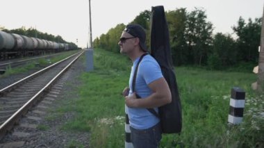 Güneş gözlüklü, gitar kılıflı adam demiryolu geçidinde duruyor.