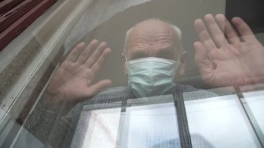 Yüz maskeli, konsantre olmuş yaşlı adam pencerede el ele tutuşuyor.