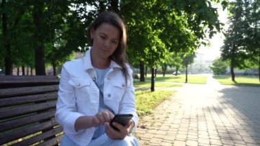 Ceketli kadın parkta telefon ekranında sörf yapıyor.