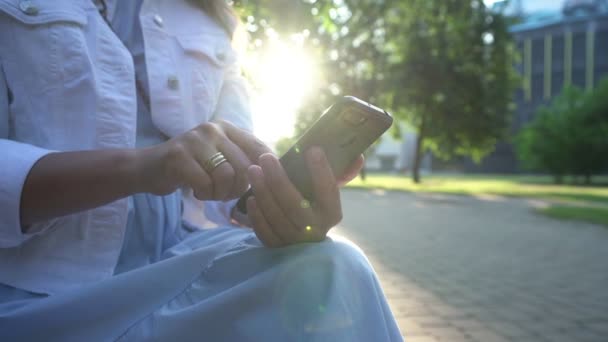 Femme en veste blanche glisse sur le téléphone dans le parc à la lumière arrière — Video