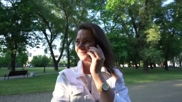 大人の女性でジャケットでウォッチトークでスマートフォンで公園 — ストック動画