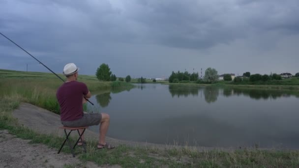 Homem senta-se na cadeira na costa do reservatório e joga vara de pesca na água — Vídeo de Stock