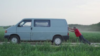 Yorgun adam eski minivanı tarla arazisinden kırsal yolda itiyor.