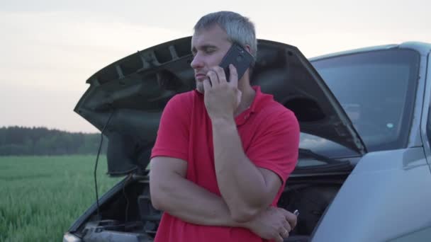 Homem fica perto de carro quebrado com capuz levantado emocionalmente falando no telefone — Vídeo de Stock