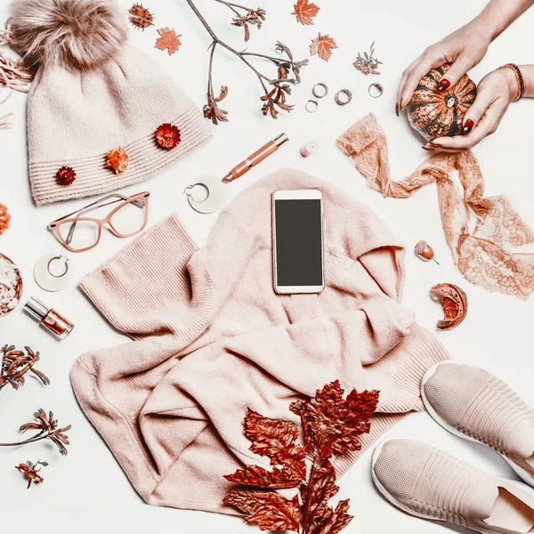 秋の女性の服に空のスクリーンスマートフォン 編んだセーター スニーカー女性の手 カボチャ 化粧品 メガネと秋の葉を構成する白いデスクトップの背景に靴 ロイヤリティフリーのストック写真