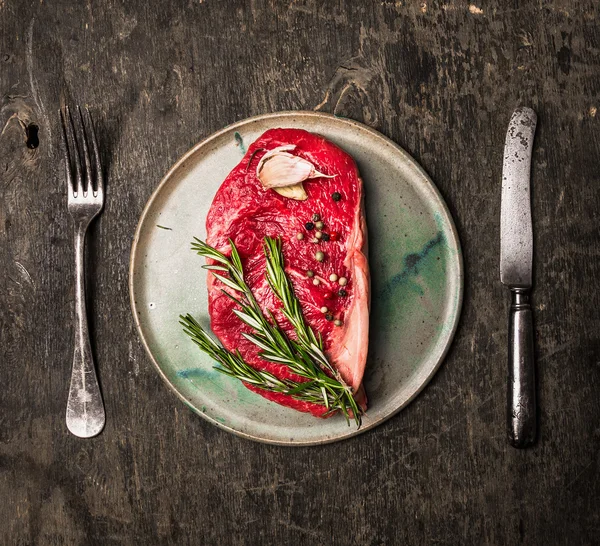 Rohes Striploin-Steak auf Teller mit Rosmarin, Knoblauch, Salz und Pfeffer auf dunklem Holzgrund, Draufsicht — Stockfoto