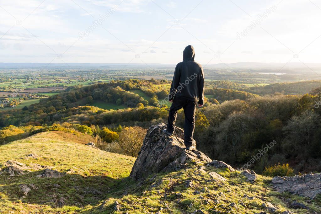 A hooded man, standing on a rocky outcrop on top of a hill, Looking out across the countryside in October. Malvern Hills, UK