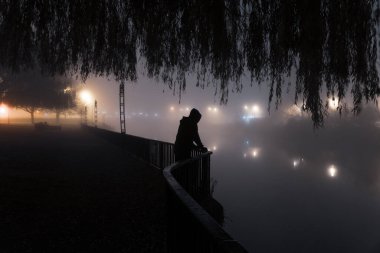Gizemli kukuletalı bir figür nehre bakıyor. Şehirde sisli bir kış gecesinde.