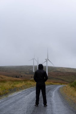 Toprak yolda duran kapüşonlu bir figür. Rüzgar türbinlerine bakıyorum. Kasvetli bir dağ yolunda. Galler. İngiltere.