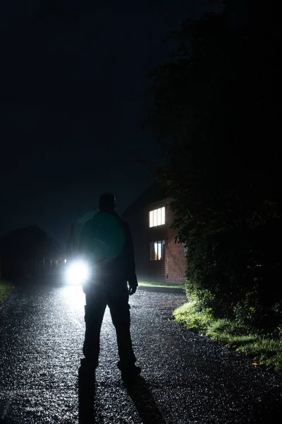 A mysterious man standing on a road, back to camera. Looking at car headlights. On a wet, dark night.