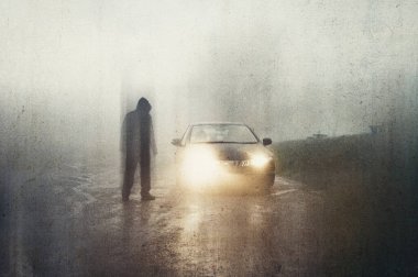 Bulanık kapüşonlu hayalet figürü. Arabanın farlarının önünde. Ürkütücü bir köy yolunda. Kırsalda sisli bir akşamda. Bir grunge, soyut düzenleme ile.