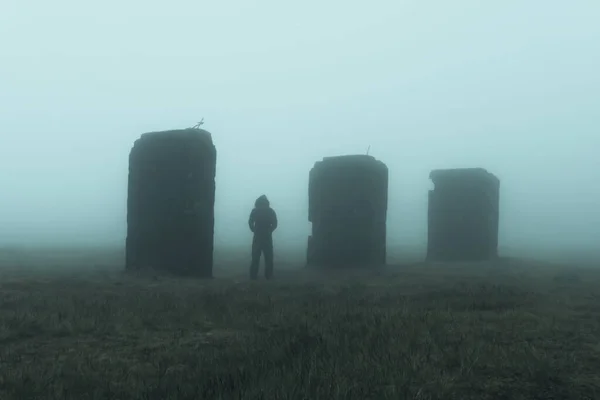 A hooded figure standing by mysterious abandoned industrial pillars, standing on moorland. On a spooky, foggy day.