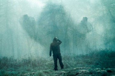 A double exposure of hooded figure looking at a scary half transparent figure appearing out of a forest. On a moody foggy winters day