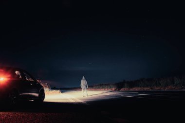 A mysterious lone figure next to a car, parked on the side of the road,  on a summers night. Standing in the middle of a road looking up at the night sky.