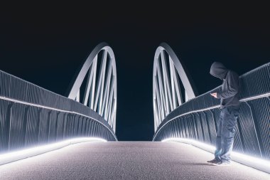 Yolda telefonuna bakan kapüşonlu gizemli bir şahıs. Geceleri parlak ışıklara karşı siluet. 