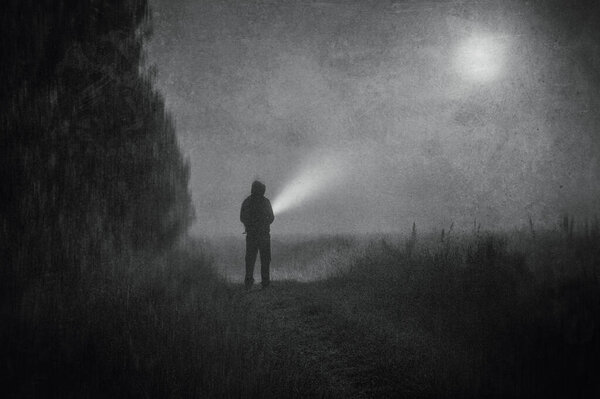 A moody atmospheric concept, of a spooky figure standing in a field with a torch on a foggy spooky night. With a grunge, vintage black and white edit