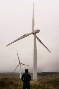 A mysterious hooded figure. Looking up at wind turbines on a mountain. On a moody winters day