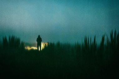 A dark scary concept. Of a mysterious figure, standing outside. Silhouetted against torch light at night. With a grunge, textured edit. 