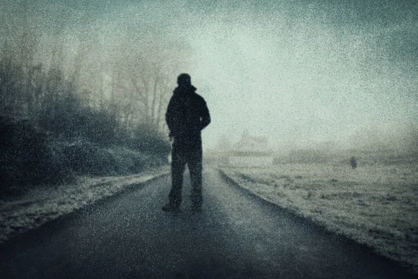 A mysterious figure standing on a road in the countryside. On an atmospheric winters day. With a grunge, textured edit.
